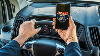 Close up of the hands of a man driver, holding phone in hand while driving, phone displays no texting while driving on screen. Very unsafe driving habit.