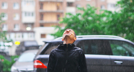 Portrait of a young man in a jacket with a hood in the rain on blurred background city street, close-up.