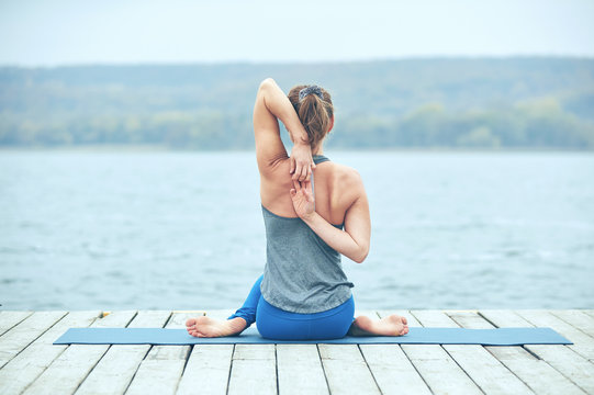 Beautiful young woman practices yoga asana Gomukhasana - Cow face pose on the wooden deck near the lake