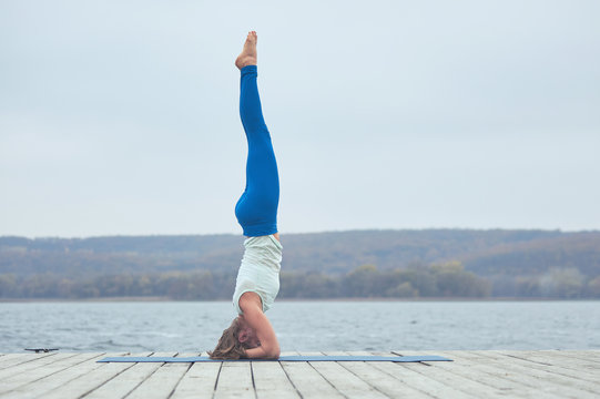 Beautiful young woman practices yoga asana Shirshasana - Headstand pose on the wooden deck near the lake