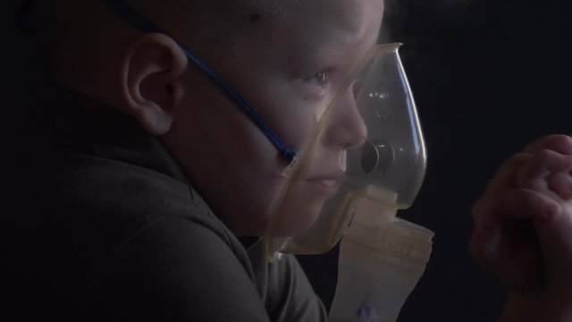 Unhappy boy with shaved head inhales vapors containing medicines for inhaler to stop cough. Child is looking information about his illness in smartphone.  Respiratory medicine. Bronchitis problem