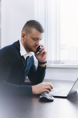 Calm serious young man uses phone while working at table in office