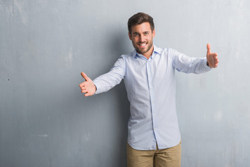 Handsome young business man over grey grunge wall wearing elegant shirt looking at the camera smiling with open arms for hug. Cheerful expression embracing happiness.