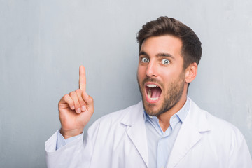 Handsome young professional man over grey grunge wall wearing white coat pointing finger up with successful idea. Exited and happy. Number one.