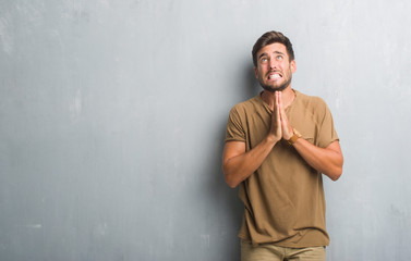 Handsome young man over grey grunge wall begging and praying with hands together with hope...