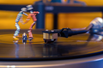 two funny small human figures, roughly welded together from resistors and transistors, dance on a rotating vinyl record in twilight; stylized as an vintage photo, focus on phonograph tonearm