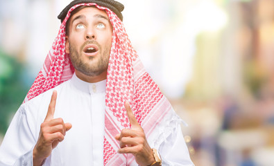 Young handsome man wearing keffiyeh over isolated background amazed and surprised looking up and pointing with fingers and raised arms.