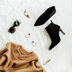 Flat lay, top view fashion concept. Trendy woman clothes and accessories on white blanket. Woolen sweater, boots, sunglasses, watch, earrings, lipstick.