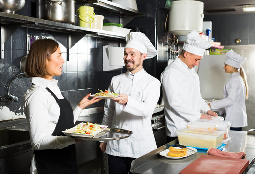 cheerful woman waiter collecting dishes from restaurant’s kitchen