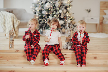 Three young children in red pajamas lie in a bed in a cozy living room and eat sweet candy on a stick. Christmas concept. Home holiday