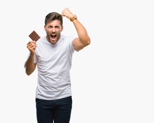 Young handsome man eating chocolate bar over isolated background annoyed and frustrated shouting with anger, crazy and yelling with raised hand, anger concept