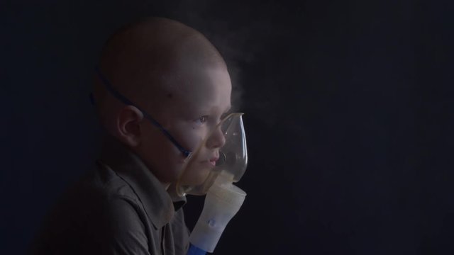 Unhappy boy with shaved head inhales vapors containing medicines for inhaler to stop cough. Child is looking information about his illness in smartphone.  Respiratory medicine. Bronchitis problem