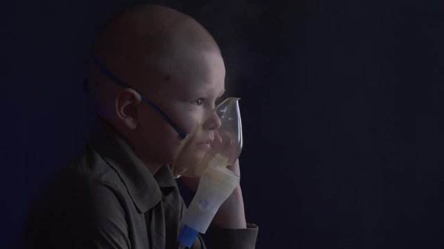 Unhappy boy with shaved head inhales vapors containing medicines for inhaler to stop cough. Child is looking information about his illness in smartphone.  Respiratory medicine. Bronchitis problem