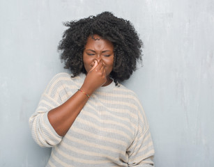 Fototapeta na wymiar Young african american plus size woman over grey grunge wall wearing a sweater smelling something stinky and disgusting, intolerable smell, holding breath with fingers on nose. Bad smells concept.