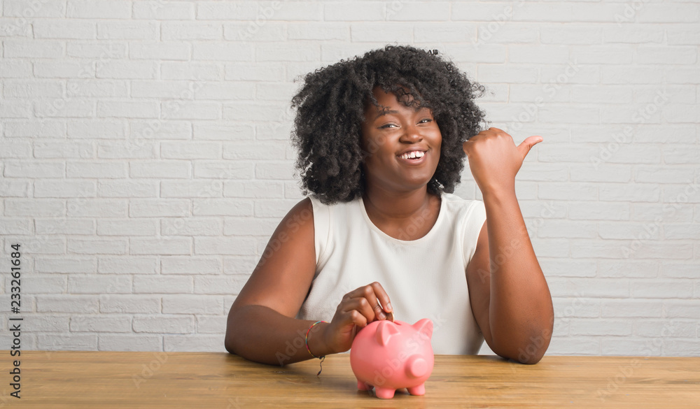 Canvas Prints Young african american woman sitting on the table holding piggy bank pointing and showing with thumb up to the side with happy face smiling