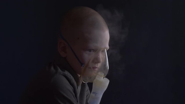 Unhappy boy with shaved head inhales vapors containing medicines for inhaler to stop cough. Child is looking information about his illness in smartphone.  Respiratory medicine. Bronchitis problem