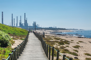 View of pedestrian walkway, with people walking and resting on the wooden platform, beside the sea, background with industry and beaches with people, blue sky, in Leca da Palmeira, Portugal
