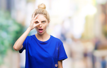 Young beautiful blonde and blue eyes woman wearing blue t-shirt over isolated background doing ok gesture shocked with surprised face, eye looking through fingers. Unbelieving expression.