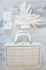 White Wicker Basket Purse with White Gloves and Handkerchief on a marble table