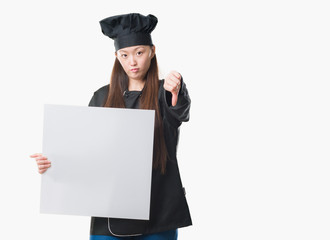 Young Chinese woman over isolated background wearing chef uniform holding banner with angry face, negative sign showing dislike with thumbs down, rejection concept