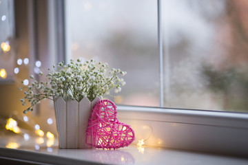 Pink wicker heart and bouquet of beautiful white gypsophila near window in the daylight, bokeh lights on background. - 233273223