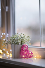 Pink wicker heart and bouquet of beautiful white gypsophila near window in the daylight, bokeh lights on background.