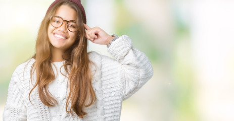 Fototapeta premium Young beautiful brunette hipster woman wearing glasses and winter hat over isolated background Smiling pointing to head with one finger, great idea or thought, good memory