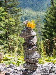 Wegzeichen aus Steinen mit gelber Blume in den Bergen am Hang unter lichten Bäumen