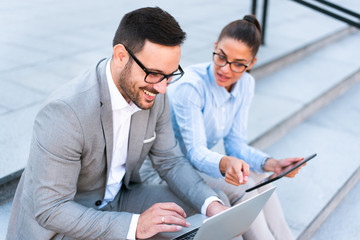 Young beautiful happy businessman and businesswoman working together outside on a laptop and tablet, smiling and talking, discussing business plans and ideas. Outdoor meeting concept.