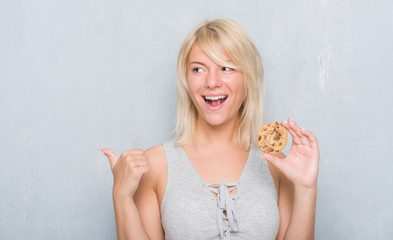 Caucasian adult woman over grey grunge wall eating chocolate cooky pointing and showing with thumb up to the side with happy face smiling