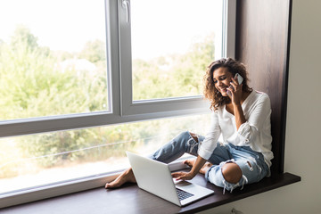 Cheerful woman mixed race playfull talking phone and sitting on windowsill with laptop computer at home