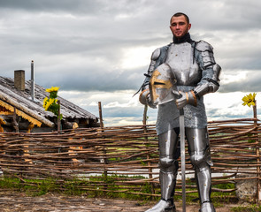 Knight in armor on the background of a wicker fence, a wooden house and the expanses of the...