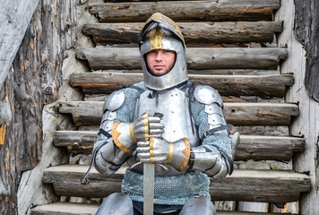 Knight in the armor on the wooden steps. Knightly armor and weapon. Semi - antique photo.