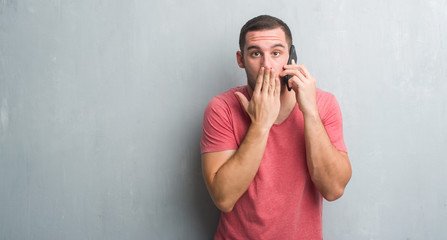 Young caucasian man over grey grunge wall talking on the phone cover mouth with hand shocked with shame for mistake, expression of fear, scared in silence, secret concept