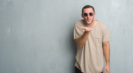 Young caucasian man over grey grunge wall wearing sunglasses looking at the camera blowing a kiss with hand on air being lovely and sexy. Love expression.