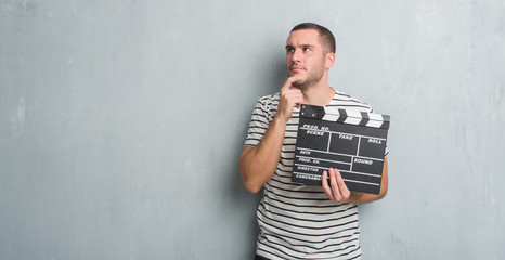 Young caucasian man over grey grunge wall holding movie clapboard serious face thinking about...