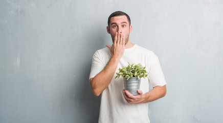 Young caucasian man over grey grunge wall holding plant pot cover mouth with hand shocked with shame for mistake, expression of fear, scared in silence, secret concept