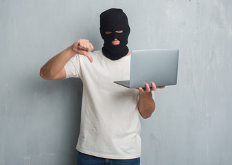 Young caucasian hacker man over grey grunge wall doing cyber attack using laptop with angry face, negative sign showing dislike with thumbs down, rejection concept