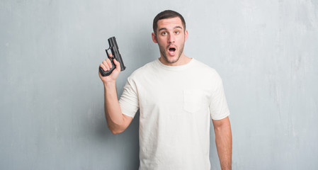 Young caucasian criminal man over grey grunge wall holding gun scared in shock with a surprise face, afraid and excited with fear expression