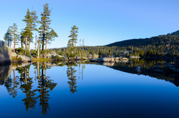 Obraz premium Pine trees, rocks, and perfect blue sky reflected in the serene, calm, and peaceful waters of an alpine lake at sunrise - Middle Velma Lake in the Desolation Wilderness near Lake Tahoe, California