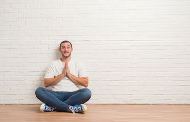 Young caucasian man sitting on the floor over white brick wall praying with hands together asking for forgiveness smiling confident.