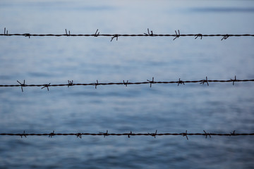 Silhouette of barbed wire fence against blurry blue water in the evening. Focused on the foreground.