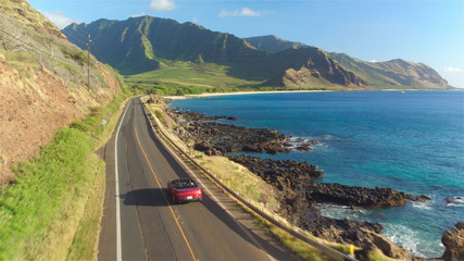 AERIAL: Couple in red convertible car driving along the beautiful coastal road - obrazy, fototapety, plakaty