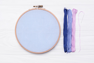 Tools for cross stitch. Threads, a hoop for embroidery, a canvas and needle on white wooden...
