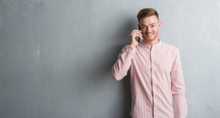 Young redhead man over grey grunge wall talking on the phone with a happy face standing and smiling...