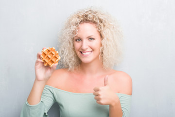 Young blonde woman over grunge grey background eating belgian waffle happy with big smile doing ok sign, thumb up with fingers, excellent sign