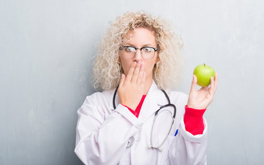 Young blonde dietitian doctor woman over grunge grey wall holding green apple cover mouth with hand...