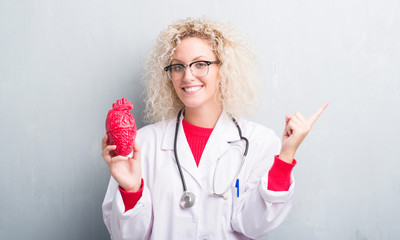 Young blonde cardiologist doctor woman over grunge grey wall holding red heart very happy pointing...