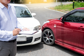 Insurance agent writing on clipboard while examining car after accident claim being assessed and processed.