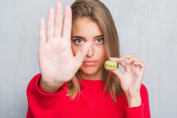 Beautiful young woman over grunge grey wall eating green macaron with open hand doing stop sign with serious and confident expression, defense gesture